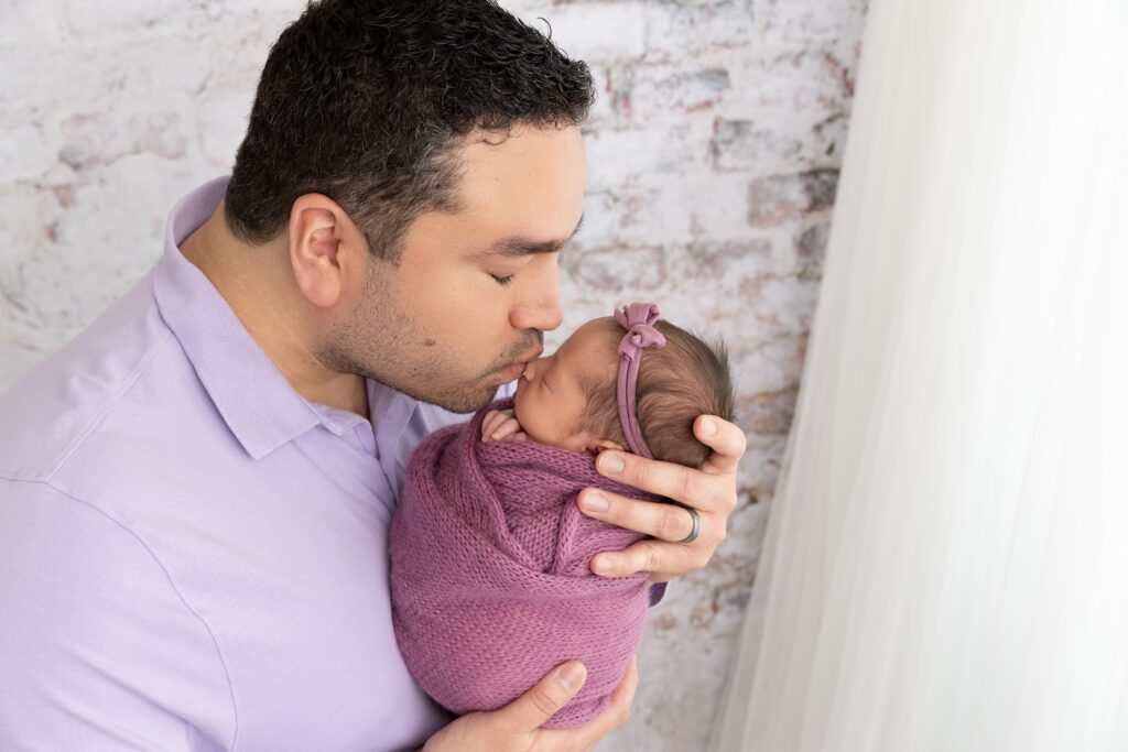 Father kissing his newborn baby wrapped in a soft purple blanket during a Phoenix newborn photography session. This heartfelt image captures the emotional connection families love in Phoenix newborn photography.