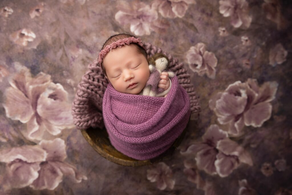 Sleeping newborn baby girl wrapped in a soft purple knit and posed in a bowl with a floral backdrop during a Phoenix newborn photography session. This artistic setup is popular with families looking for Scottsdale newborn photos and Arizona newborn sessions.