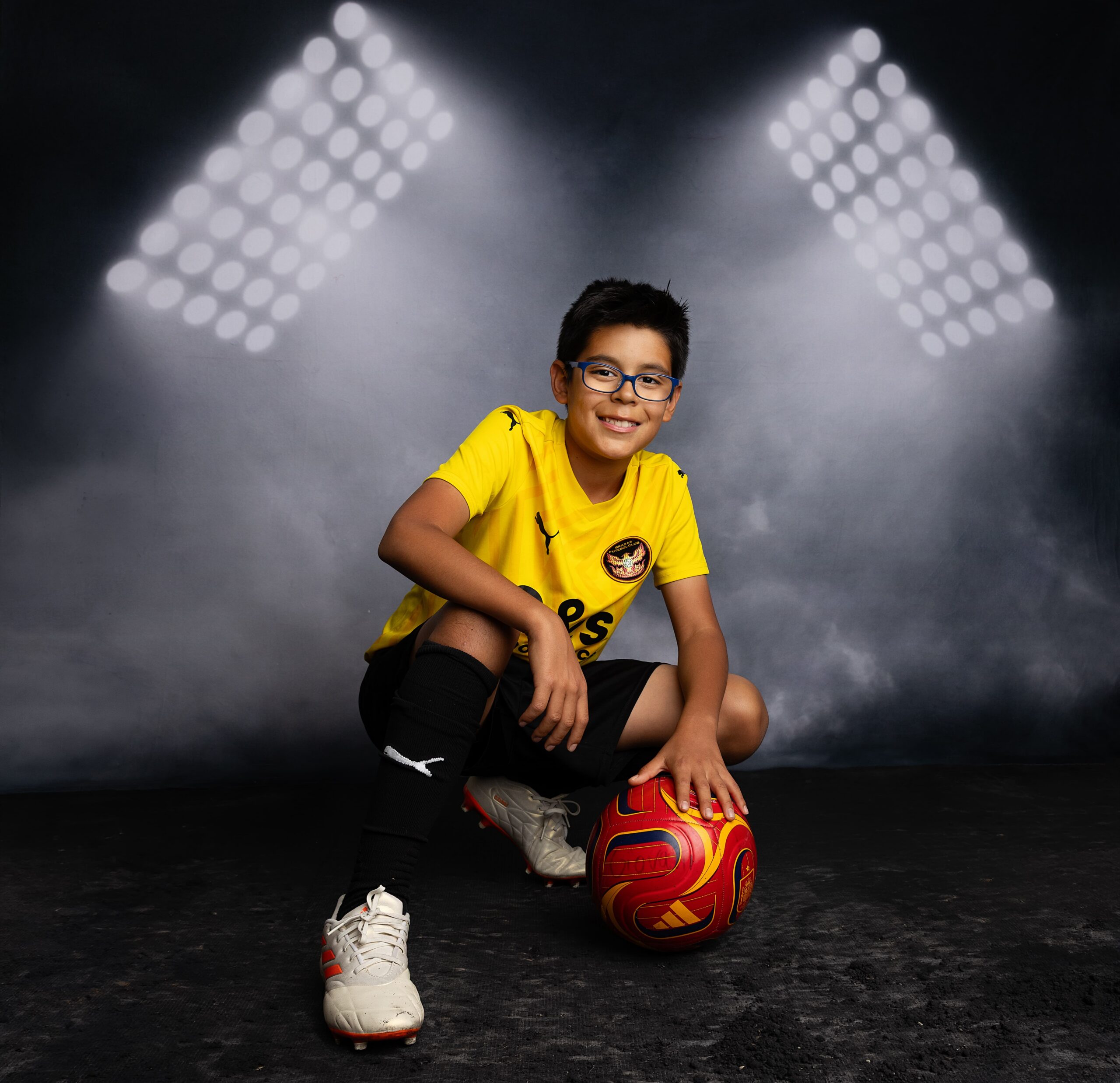 Youth soccer player in yellow jersey posing with ball in a Phoenix studio sports portrait with stadium-style lighting.